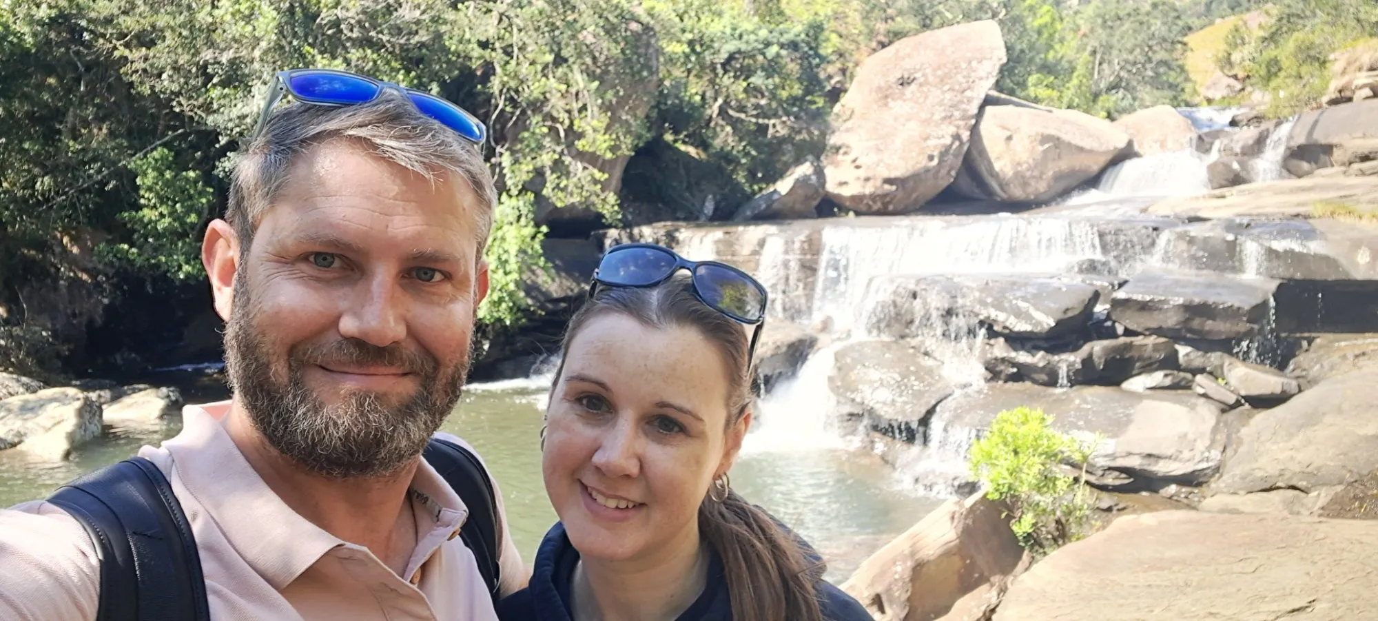 Nadine and Egan smiling in a selfie in front of a waterfall cascading over large rocks